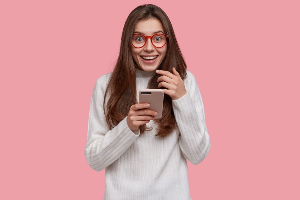 Técnicas de persuasão para aumentar a taxa de abertura dos emails - Jovem usando óculos vermelho com celular na mão e expressão de surpresa.