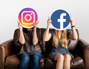Descubra qual é a diferença entre impulsionamento e tráfego pago - Duas mulheres sentadas em um sofá marrom segurando um círculo com o ícone das redes sociais Instagram e Facebook na frente do rosto.