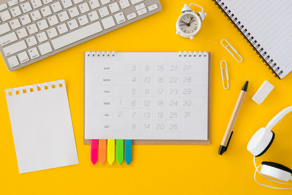 Como é o dia a dia de um Copywriter Profissional? - Notebook, calendário, caderno, caneta, relógio e clipes espalhados sobre um fundo amarelo ouro.