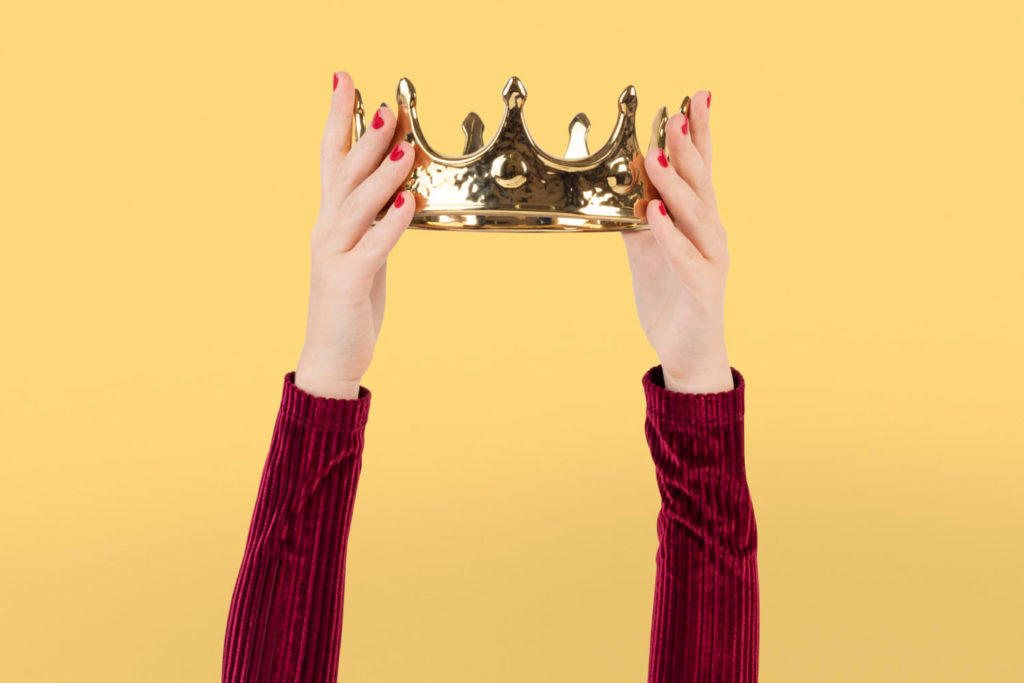 Os 10 melhores Copywriters do Brasil (Bettina está na lista) - Mãos femininas erguendo uma coroa sobre um fundo amarelo