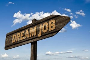 3 razões do mercado de trabalho tradicional remunerar menos que o mercado digital - Placa de madeira escrita "Dream Job" apontando para a transversal sobre um fundo de um céu azul com nuvens brancas